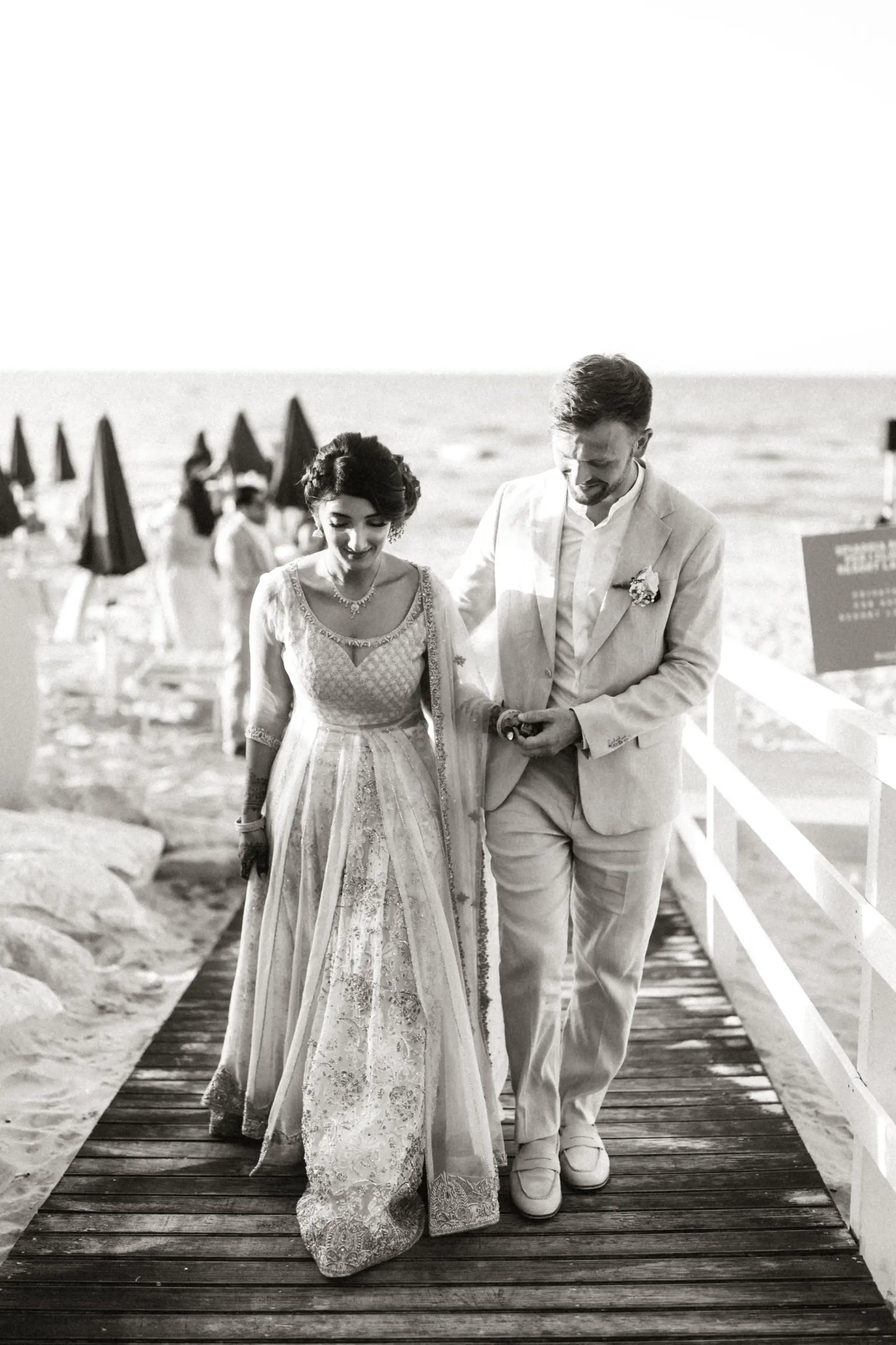 Bride and groom walking hand in hand along a boardwalk at their beach wedding in Alcamo Marina, Sicily, dressed in traditional South Asian and Mediterranean attire
